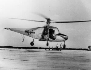 A Higgins helicopter flies over a flat surface, with two men looking on from the ground, at a distance.
